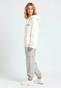 Felpa crema con cappuccio e coulisse regolabile, tasca anteriore e scritte rosse; abbinata a pantaloni cargo grigi chiari e sneakers bianche.