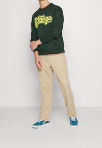 Zelená mikina s žlutým textovým designem, béžové kordové kalhoty a azurové tenisky s bílými detaily. Strukturovaná látka, klasický střih.