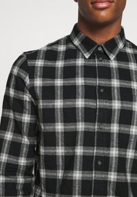 Camicia in flanella a quadri nera e grigia con colletto button-down, dotata di tasca e tessuto testurizzato per il calore. I bottoni sono neri.