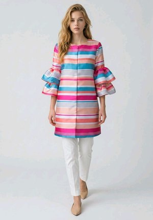 Mujer con abrigo de rayas coloridas con mangas con volantes, pantalones blancos y zapatos beige, de pie frente a un fondo claro liso.