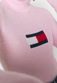 Růžový žebrovaný rolák se pletenou nášivkou loga ve navy, červené a bílé, umístěnou uprostřed přední části.