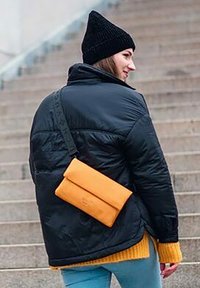 Černá zateplená bunda s vysokým límcem, zkombinovaná se žlutým svetrem a oranžovou kabelkou přes rameno, doplněná černou čepicí.