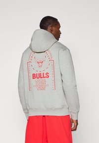 Grå hættetrøje med et rygdesign, der har en rød grafik af en tyr og tekst, der henviser til "Chicago Bulls" og detaljer om basketballdivisionen.