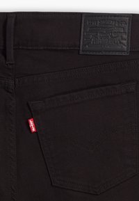Vaqueros de denim negro con un acabado texturizado, que presentan una etiqueta de cuero en la cinturilla y una etiqueta de marca roja en el bolsillo trasero.