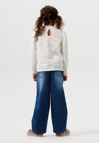 White long-sleeve top with eyelet pattern, high neck, and back keyhole. Paired with wide-leg blue denim jeans. Multi-colored sandals.