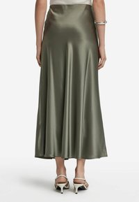 Olivegrüner, satinierter Maxi-Rock mit glatter Textur, ausgestelltem Design und seitlichem Schlitz. Getragen mit weißen High Heels.