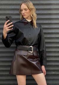 Camisa de cuero sintético negra con un cuello burdeos, combinada con una mini falda de cuero marrón que cuenta con un bolsillo frontal y un gran cinturón de hebilla.