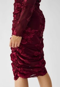 Burgunderfarbenes, tailliertes Kleid mit floraler Stickerei, gerafftem Textil, durchsichtigen Ärmeln und gewelltem Saum. Elegante, figurbetonte Silhouette.