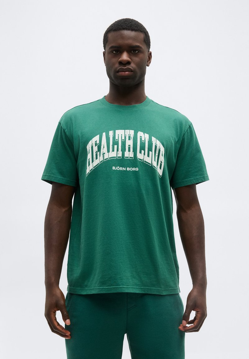 Νέος άνδρας που κοιτάζει μπροστά, φορούντας πράσινο T-shirt με την επιγραφή «HEALTH CLUB» και ταιριαστό πράσινο παντελόνι, μπροστά σε απλό ανοιχτό φόντο.