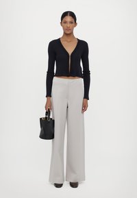 Svart ribbestrikket kort cardigan med dyp V-hals, kombinert med lysegrå vide bukser. Modellen holder en liten svart håndveske.
