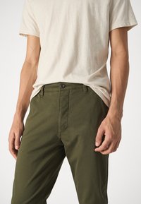 Chinos en coton vert olive avec une coupe droite, présentant une fermeture éclair à l'avant, deux poches latérales et une fermeture à un bouton à la taille.