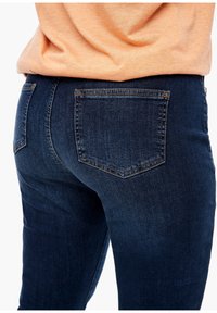 Jeans bleu marine avec une texture lisse et un léger éclat, présentant deux poches arrière et des détails de couture contrastants.