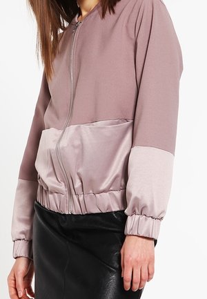 Femme portant une veste bomber mauve zippée avec des poignets et un ourlet élastiques, associée à une jupe en cuir noire.
