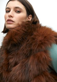 Hnědý umělý kožich s dlouhými, jemnými vlákny, které mají luxusní texturu. Nositý přes tyrkysový oděv. Detailní záběr zdůrazňuje detaily.