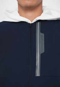 Kratak prikaz osobe koja nosi tamnoplavu jaknu na zatvarač s bijelom kapuljačom i vertikalnim reflektirajućim EA7 Emporio Armani logotipom na prsima.