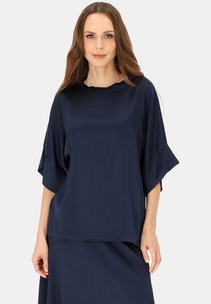 Kobieta z długimi brązowymi włosami ubrana w luźny granatowy top z szerokimi rękawami i pasującą spódnicę na jednolitym jasnym tle.