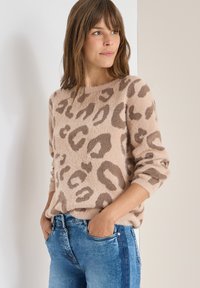 Jasnobrązowy sweter w panterkę wykonany z miękkiego, puszystego materiału. O okrągłym dekolcie, z długimi rękawami i luźnym krojem. Połączony z niebieskimi dżinsami.