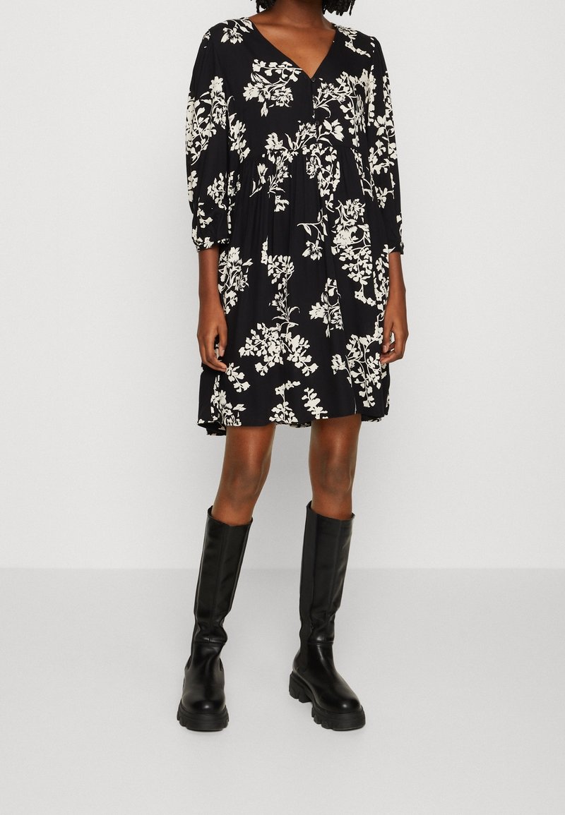 Robe noire à fleurs avec un décolleté en V, des manches trois-quarts et un corsage froncé, associée à des bottes en cuir noires montantes.