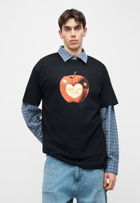 Νέος άνδρας που φοράει μαύρο T-shirt με κόκκινο μήλο και καρδιά πάνω από μπλε καρό πουκάμισο με κουμπιά και ανοιχτό μπλε τζιν.