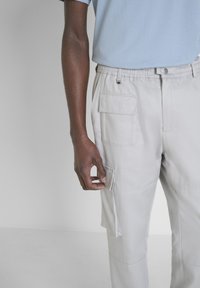 Pantalons cargo gris clair avec plusieurs poches, taille élastique et texture brossée. Main tenant un détail de poche latérale.