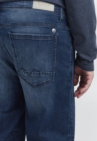 Jeans od denima u tamnoplavoj boji s klasičnim krojem. Sadrži označavanje na pojasu i stražnji džep s trokutastim šavom.
