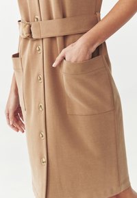 Robe beige à boutons sur le devant, avec ceinture et deux grandes poches avant, une main partiellement dans la poche droite.