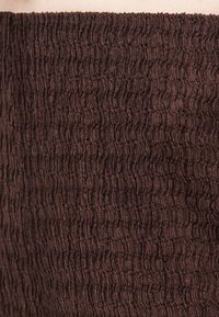 Gros plan d’un tissu marron foncé avec une texture smockée serrée et ondulée et un détail élastique froncé.