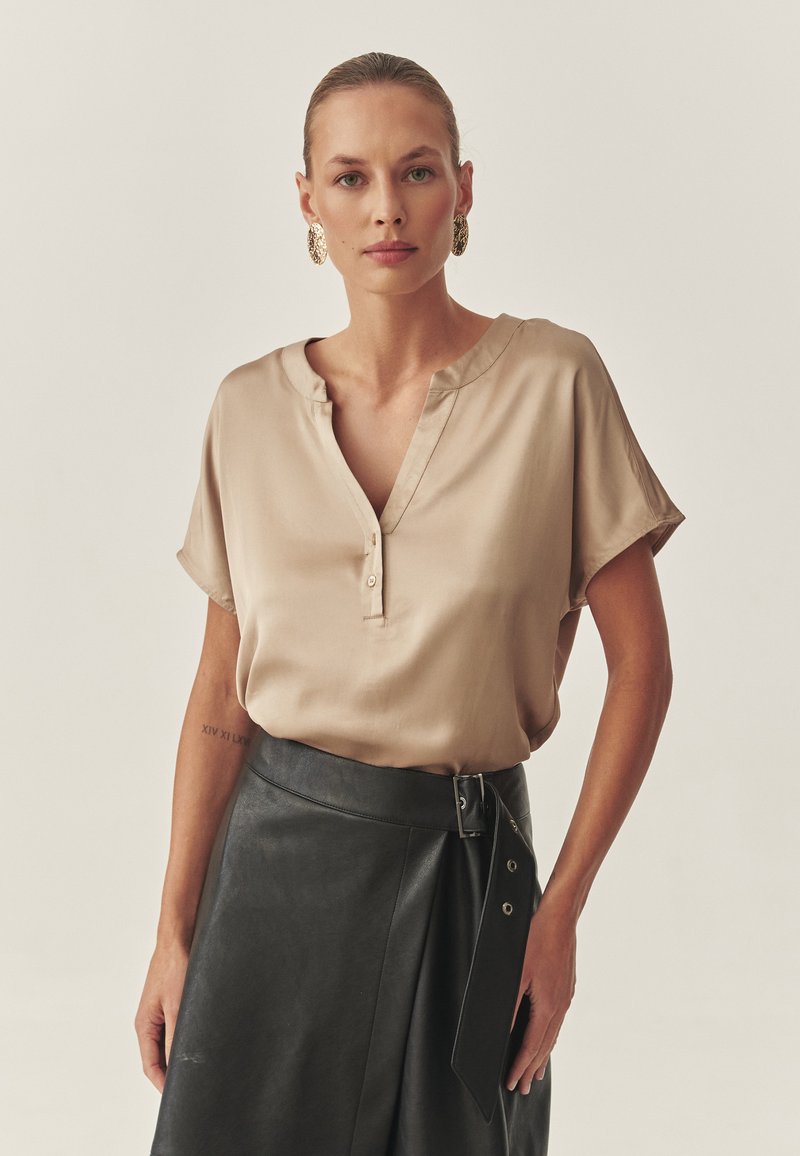 Beige sidenblus med V-ringning och korta ärmar. Bärs tillsammans med en svart läderkjol med bälte i midjan och asymmetrisk fickdetalj.