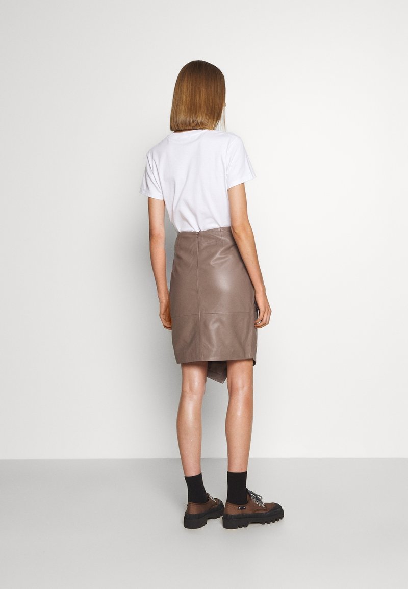 Kvinde stående med ryggen vendt mod os iført en hvid t-shirt, taupe lædernederdel, sorte sokker og brune snørestøvler mod en ensfarvet baggrund.