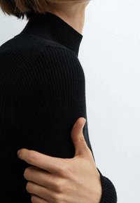 Pull noir en maille côtelée avec un design ajusté, un tissu texturé et un col haut. Gros plan d'une main reposant sur le bras.