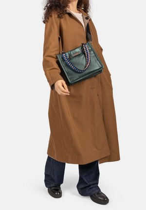 Persoon die een lange bruine jas draagt, donkere spijkerbroek, zwarte schoenen en een donkergroene handtas met blauw en roze gevlochten handvatten.