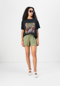 Femme portant un t-shirt graphique noir, un short vert olive à cordon de serrage, des chaussures blanches et des lunettes de soleil surdimensionnées, sur un fond blanc uni.