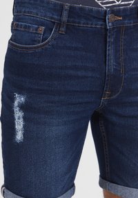 Džínové šortky tmavomodrej farby s opotrebovanými detailmi a zavinutými lemami, vybavené štandardnými vreckami a kontrastným prešívaním.
