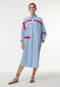 Blauwe shirtjurk met rode gerimpelde accenten, knoopsluiting aan de voorkant, lange mouwen en zakken. De stof lijkt glad met een gestructureerd ontwerp.