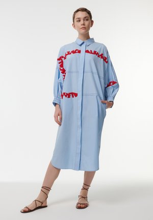 Robe chemise bleue avec des accents froncés rouges, fermeture à boutons sur le devant, manches longues et poches. Le tissu semble lisse avec un design structuré.