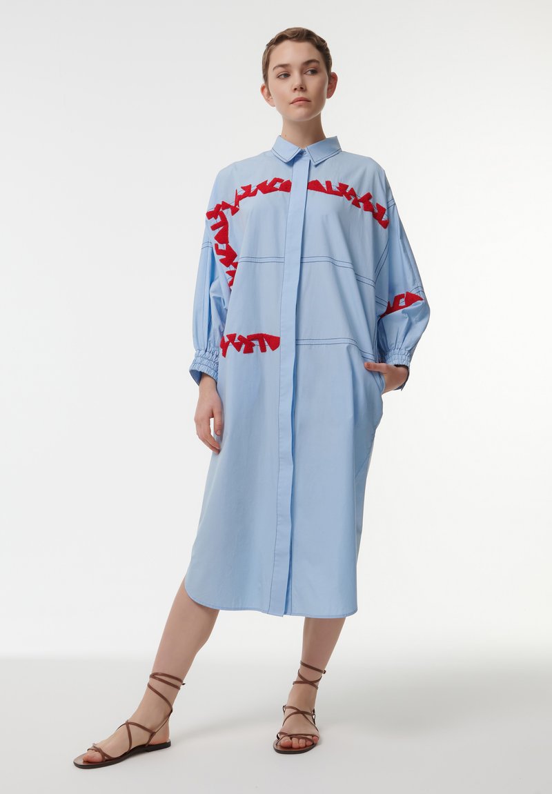 Blauwe shirtjurk met rode gerimpelde accenten, knoopsluiting aan de voorkant, lange mouwen en zakken. De stof lijkt glad met een gestructureerd ontwerp.