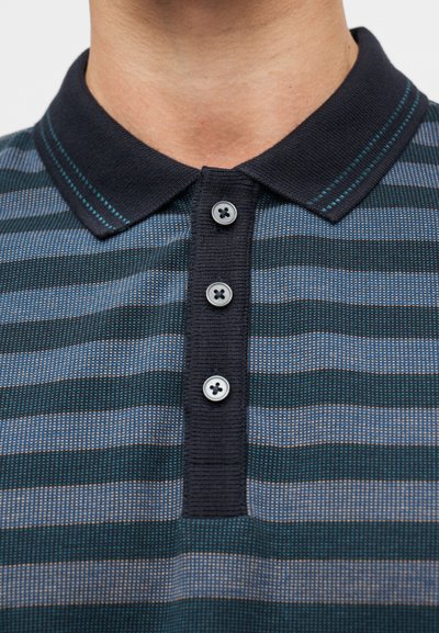 Πουκάμισο πόλο με ρίγες που παρουσιάζει οριζόντια μοτίβα σε σκούρο πράσινο, μπλε και κρεμ, με γιακαδάκι σε ναυτικό μπλε, τέσσερα κουμπιά και τυρκουάζ ραφές ως διακοσμητικά στοιχεία.