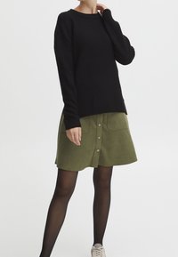 Czarny sweter z fakturą, z okrągłym dekoltem, zestawiony z zieloną spódnicą z przodu zapinaną na guziki oraz z dwoma kieszeniami. Modelka ma na sobie czarne rajstopy i trampki.