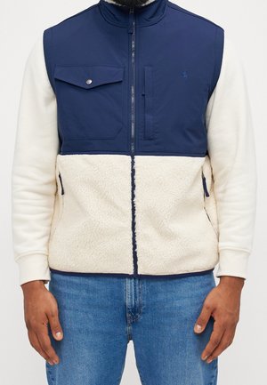 Mand iført en to-farvet marineblå og cremefarvet vest med lynlås, fleece-tekstur og forlommer over en hvid langærmet skjorte og blå jeans.