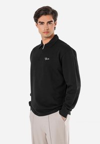 Sort langærmet polo shirt med krave og en halvlynlås, med et lille broderet logo på brystet. Blød stofstruktur.