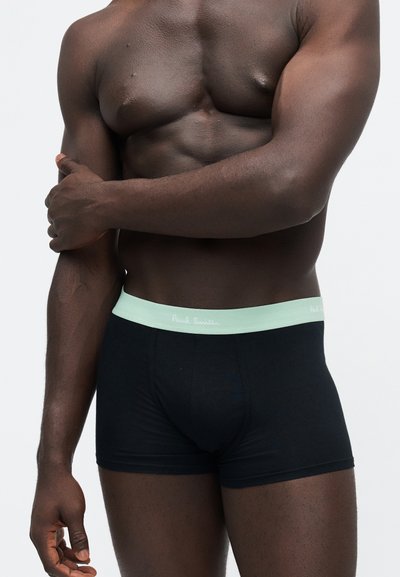 Homme musclé à la peau foncée portant un boxer noir avec une ceinture vert clair, debout devant un fond clair uni.