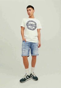 Jovem vestido com uma t-shirt branca com estampado, calções de ganga azuis claros rasgados, meias brancas e ténis pretos e brancos, posando em frente a um fundo simples.