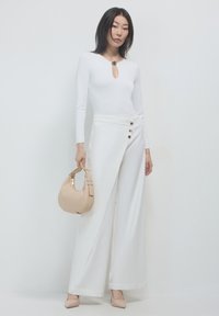 Bodysuit blanc à manches longues avec décolleté en trou de serrure, associé à un pantalon large blanc avec des accents de boutons et un sac à main beige.