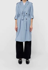 Jasnoniebieski płaszcz trencz o dwurzędowym kroju, zawiązywany paskiem w talii, podwiniętymi rękawami i bocznymi kieszeniami, zestawiony z czarnymi spodniami o szerokich nogawkach.