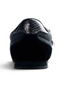 Scarpa loafer nera con tacco cap altamente lucido e corpo in suede liscio, vista da dietro su una superficie bianca.