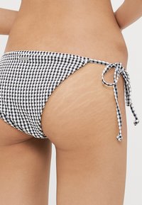 Bas de bikini en vichy noir et blanc avec des fermetures à nouer sur les côtés. Caractéristiques d'un tissu texturé et d'une coupe taille basse. Peau visible avec des vergetures.