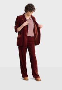 Aksamitny burgundowy garnitur z szerokimi spodniami i marynarką. Pod spodem biała koszula w kratę. Uzupełnieniem stylizacji są masywne drewniane chodaki.