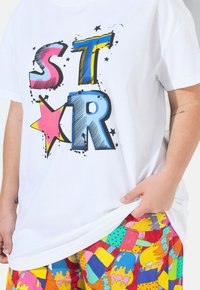 Camiseta blanca de algodón con letras de colores en bloques que dicen "STR" y estrellas. Combinada con pantalones cortos estampados en colores brillantes y motivos de helados.