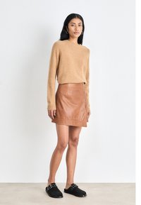 Pull en maille beige, jupe mini en cuir marron avec fermeture éclair argentée, et chaussures noires à enfiler avec des accents en métal. Fond neutre.