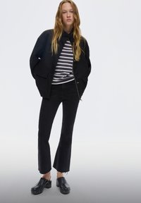 Mladá žena s dlouhými blond vlasy, oblékající černou bundu, černé zkrácené rozšířené džíny, černé klobouky na podpatku a tričko s černo-bílým proužkem.